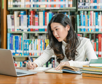 Student in a library looking at a laptop screen while writing notes with in the background various books on a book shelf.