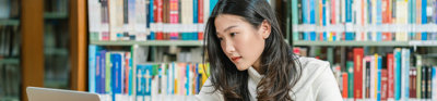 Student in a library looking at a laptop screen while writing notes with in the background various books on a book shelf.