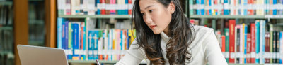 Student in a library looking at a laptop screen while writing notes with in the background various books on a book shelf.