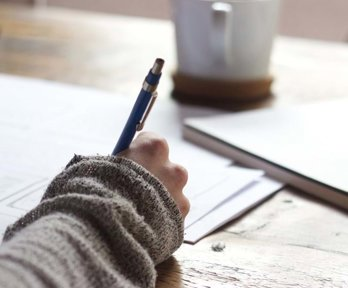 A hand holding a pen and writing at a wooden desk. A cup of coffee is in the background.