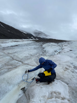 A researcher crouching on a glacier using measuring equipment, with mountains in the background.