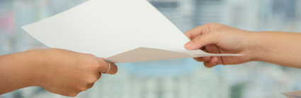 Two people hold a sheet of blank paper as one passes it to the other. Only their hands can be seen and the background is blurred.