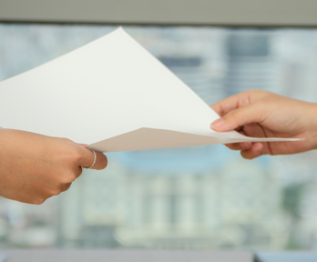 Two people hold a sheet of blank paper as one passes it to the other. Only their hands can be seen and the background is blurred.
