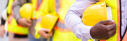 Chest-level close up of people lined up while holding hard constructions helmets and wearing yellow high vis clothing.