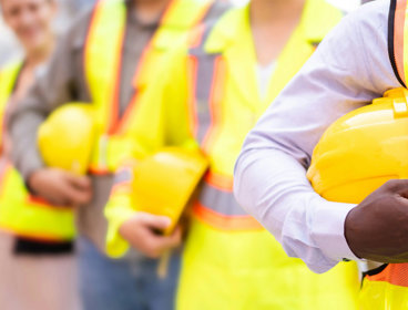 Chest-level close up of people lined up while holding hard constructions helmets and wearing yellow high vis clothing.