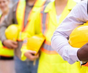 Chest-level close up of people lined up while holding hard constructions helmets and wearing yellow high vis clothing.