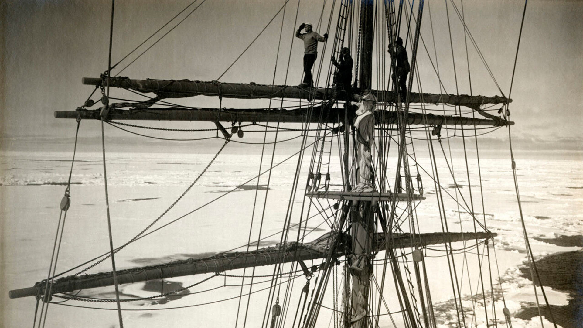 Black and white photograph of the Terra Nova ship, with people standing and sitting on the mast of the ship.