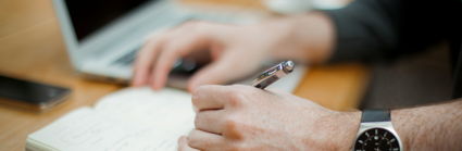 A person uses a pen pen to write on a notpad. Their phone and laptop sit next to the notepad on the wooden desk 