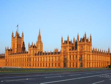 Photograph of the houses of parliament taken from the adjacent bridge with a double-decker bus crossing the bridge. 