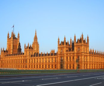 Photograph of the houses of parliament taken from the adjacent bridge with a double-decker bus crossing the bridge. 
