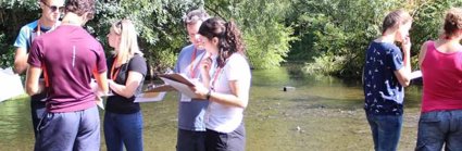 A group of teachers are standing in a shallow river, looking at clipboards