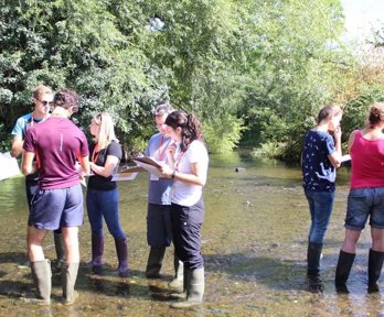 A group of teachers are standing in a shallow river, looking at clipboards