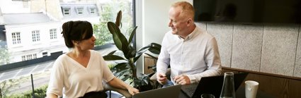 Two professionals having a conversation in an office with laptops in front of them