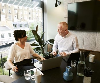 Two professionals having a conversation in an office with laptops in front of them