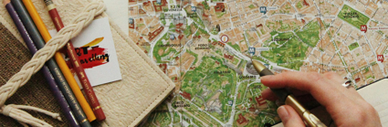 A person uses a pen to point to a location on a map. a notebook and pencils also sit on the map
