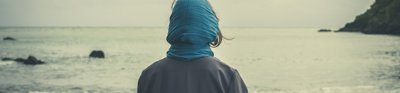 A person with a blue scarf and grey jacket looks out onto the sea