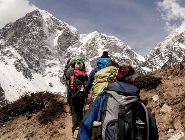 A group of people walk up mountainous terrain. They each carry large bags and walking equipment.