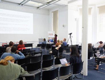 Several people sit in a modern, well-lit presentation room listening to a presenter talk.