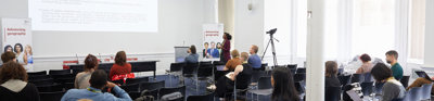 Several people sit in a modern, well-lit presentation room listening to a presenter talk.