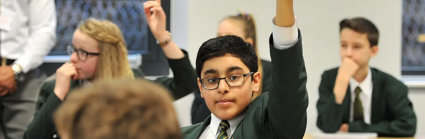 A young child wearing a school uniform sits in a classroom with their hand raised above their head to indicate they want to ask a question.