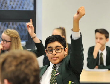 A young child wearing a school uniform sits in a classroom with their hand raised above their head to indicate they want to ask a question.