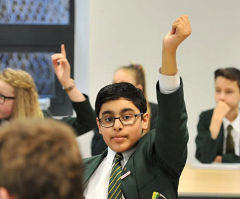 A young child wearing a school uniform sits in a classroom with their hand raised above their head to indicate they want to ask a question.