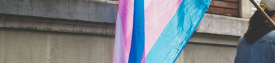 A blue, pink, and white trans flag hangs via a flag pole is held by a person faced away from the viewer and partially cut out of frame.