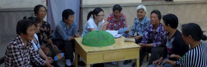 Group of people sat outside around a table. Some of them are holding pieces of paper and conversing. 