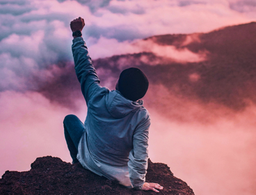 Man sitting on mountain cliff facing white clouds, with one hand in the air