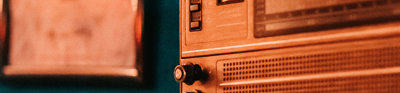 A brown retro radio in front of a dark blue wall. A blurred out framed image hangs on the wall behind the radio on the left hand side.