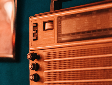 A copper coloured retro radio sits on front of a dark blue wall. A framed image hangs on the wall behind the radio on the left hand side but its too blurred out to be identified in greater detail.