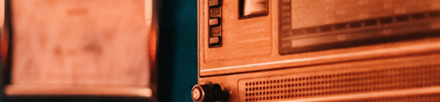 A copper coloured retro radio sits on front of a dark blue wall. A framed image hangs on the wall behind the radio on the left hand side but its too blurred out to be identified in greater detail.