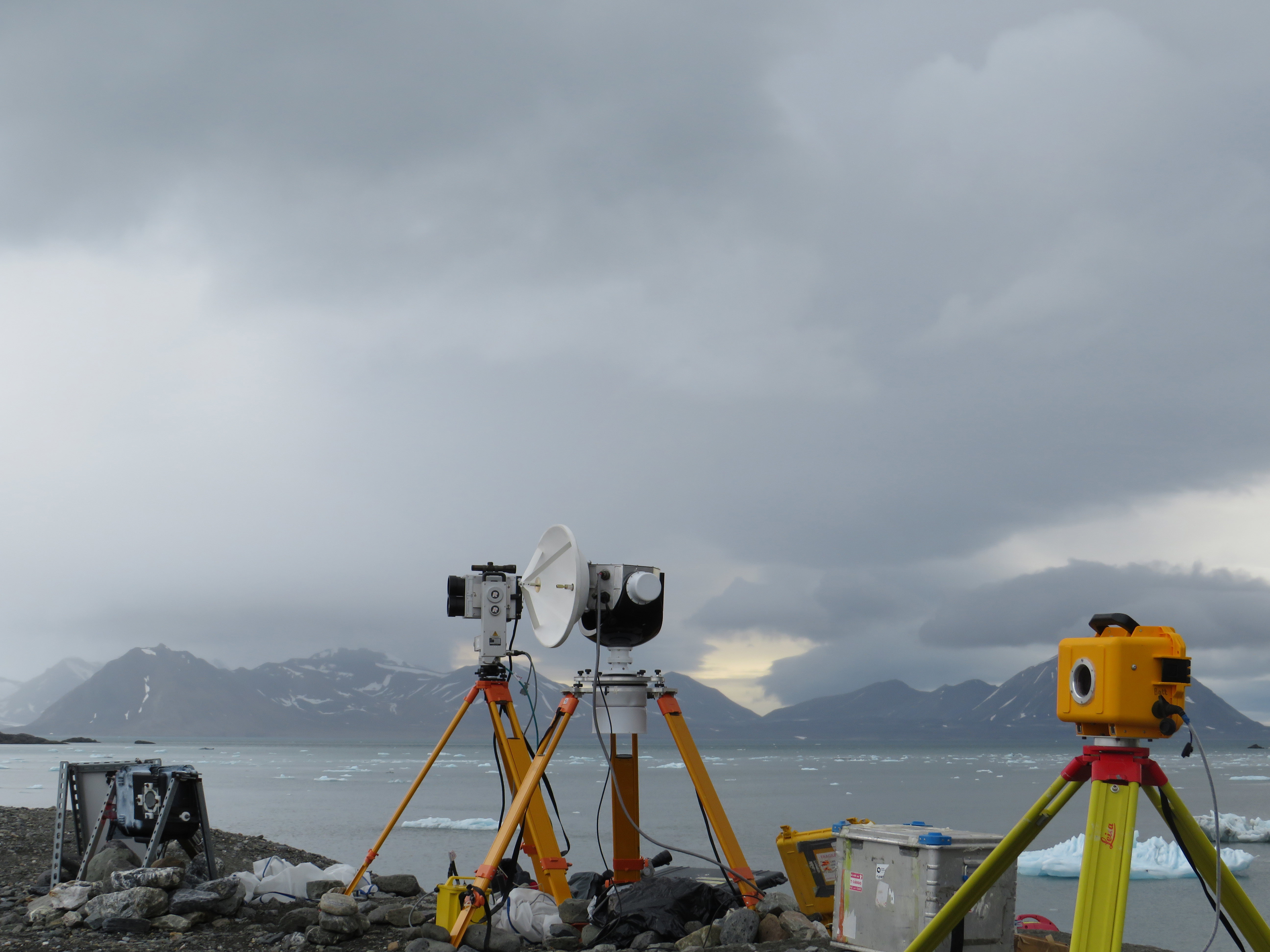 Three imaging cameras set up on tripods next to a lake, with mountains in the background. 