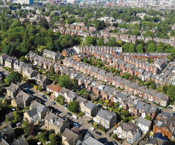 An aerial view of a residential area, with orange houses with grey roofs and small green gardens. The suburban area stretches off into the distance, with a few high rise tower blocks also in the view.