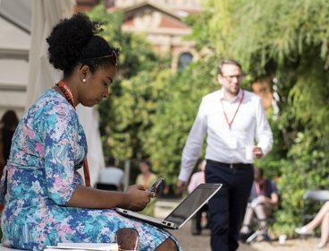 A Black woman in a blue floral dress sits in the Society's garden with her laptop on her lap as other people sit and walk nearby.
