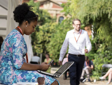 A person in a blue floral dress sits in the Society's garden with a laptop on their lap as other people sit and walk nearby.