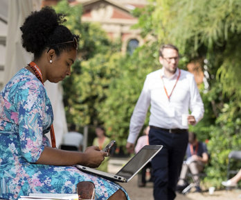 A person in a blue floral dress sits in the Society's garden with a laptop on their lap as other people sit and walk nearby.