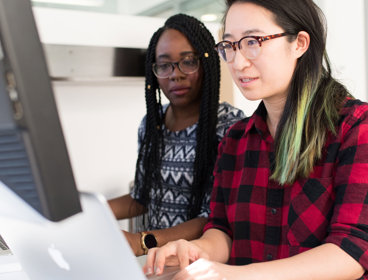 Two people sit next to each other at a desk. one uses the computer whist the other watches them. 