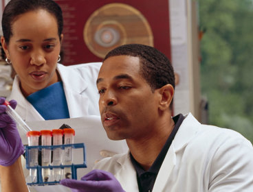 One researcher in a lab coat and disposable gloves holds a test tube rack whilst placing a test tube inside. A second researcher stands beside the first watching. 