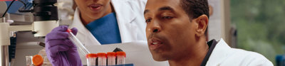 One researcher in a lab coat and disposable gloves holds a test tube rack whilst placing a test tube inside. A second researcher stands beside the first watching. 