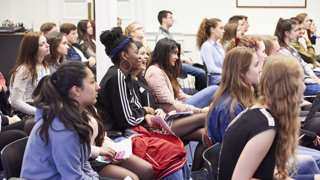 An audience on students of mixed gender and ethnicity listening to a presentation, some are smiling or laughing.