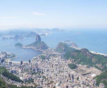 A view over Rio de Janeiro showing a sprawling city located in the valley between green wooded hills, finishing at the ocean
