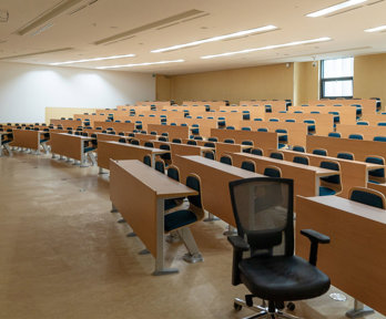 Empty lecture hall the lights on and a single office chair on the opposite side of the hall from the the lecture podium.