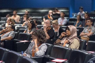 Audience of mixed gender, age and ethnicity sitting in a lecture theatre looking thoughtful while watching a presentation.