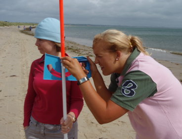 Two female students on a sandy beach, one is holding an ranging pole and the other is using an instrument to measure the height of dunes in the distance