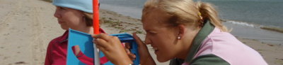 Two female students on a sandy beach, one is holding an ranging pole and the other is using an instrument to measure the height of dunes in the distance