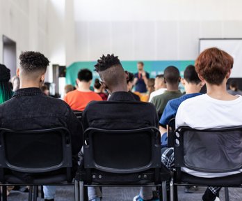 Students sitting in a classroom watching a teacher, viewed from behind