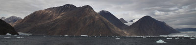 Sea with ice floating in it, with in the background a mountain range with dark grey clouds above it. 