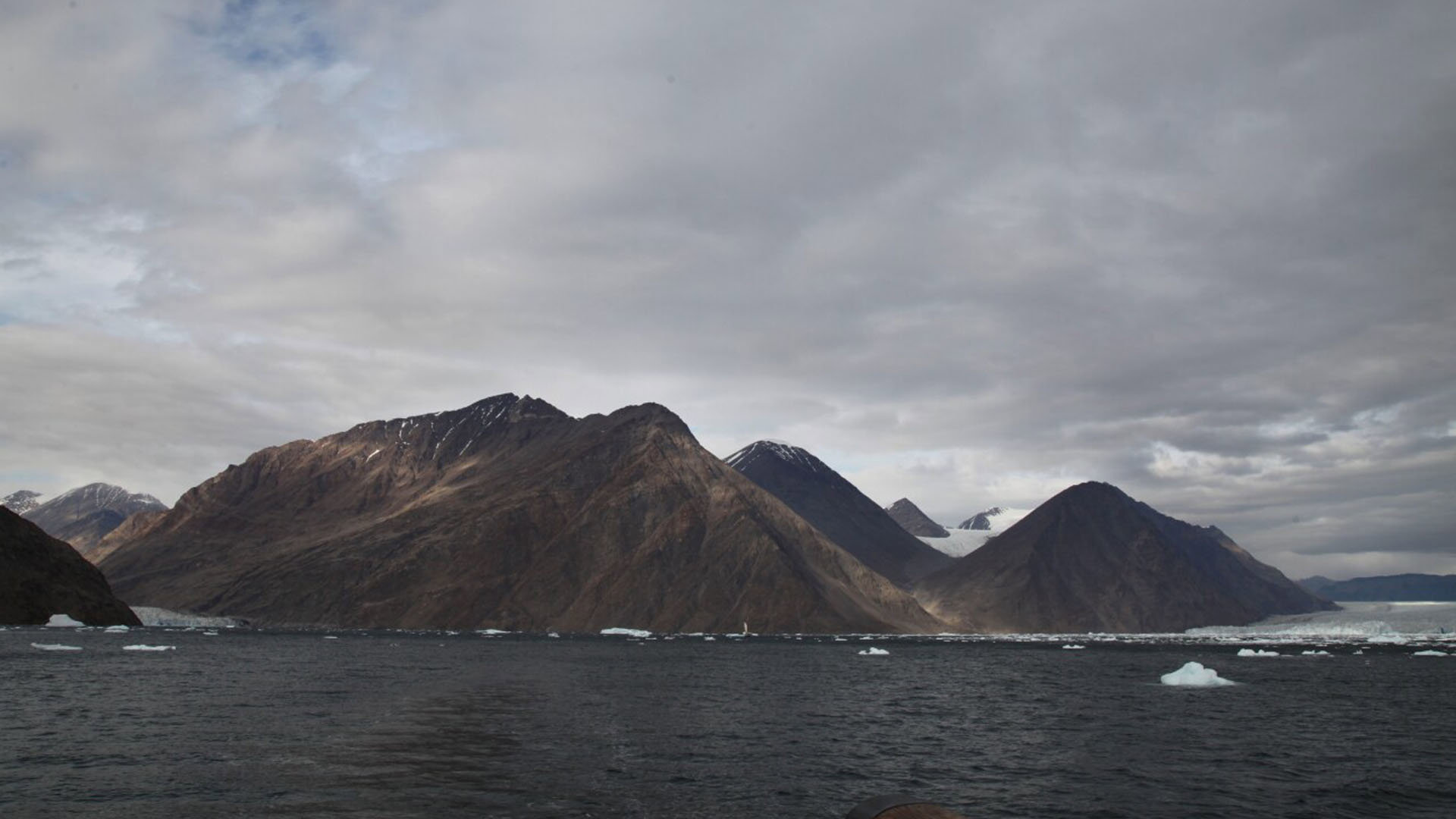 Sea with ice floating in it, with in the background a mountain range with dark grey clouds above it. 