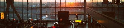 The sun rise casts light on Heathrow airport tarmac. Planes can be seen stationed on the tarmac through a large window. The dimly lit interior of the airport can be seen before the window, in the foregroun.